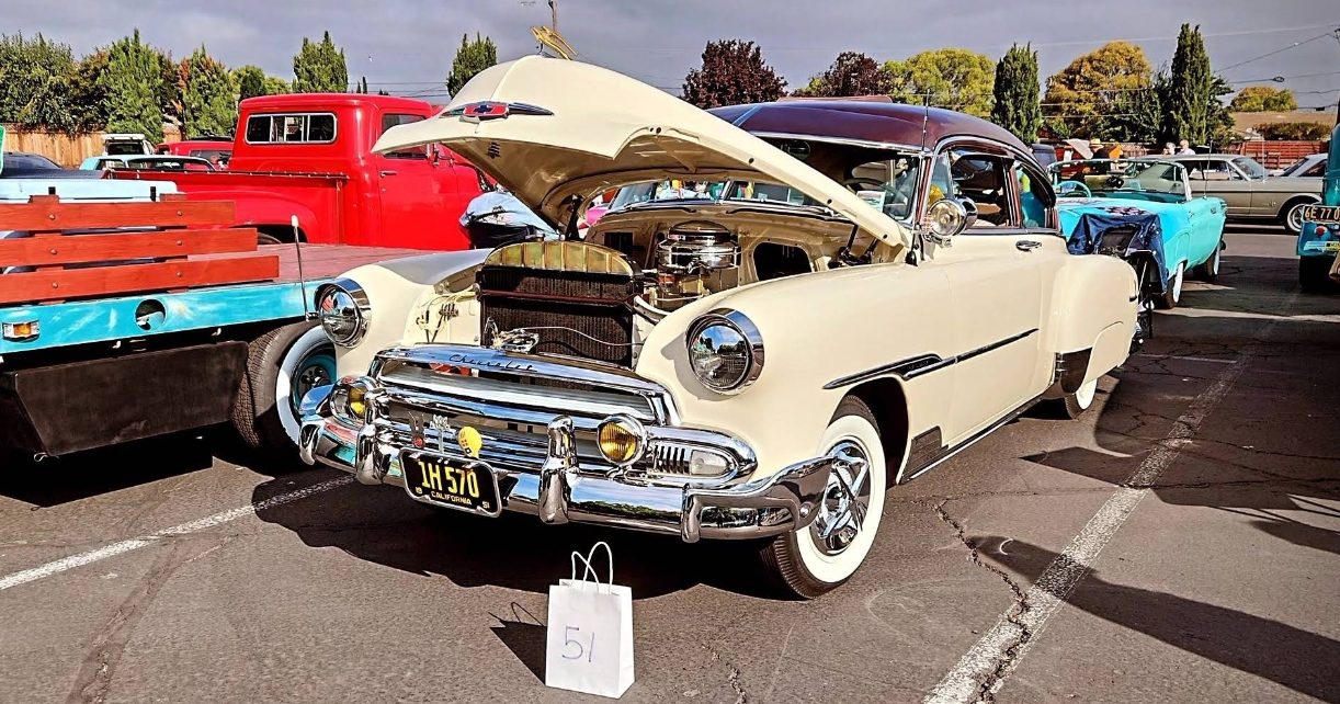 A restored cream-colored 1950s Chevrolet with its hood open at a classic car show, parked among other vintage cars and a red pickup truck.