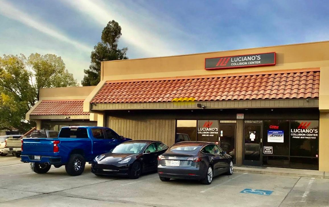 The exterior of Luciano's Collision Center in Milpitas, with two black Tesla sedans and a blue pickup truck parked in front of the storefront.